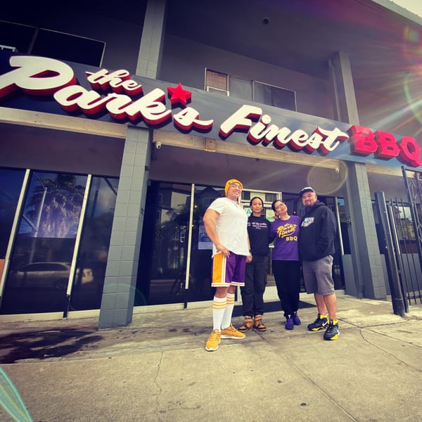 Four people stand smiling in front of The Park's Finest BBQ restaurant, with a bright sun flare.