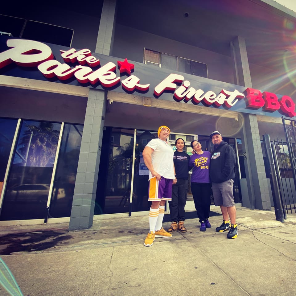 Four people stand smiling in front of The Park's Finest BBQ restaurant, with a bright sun flare.
