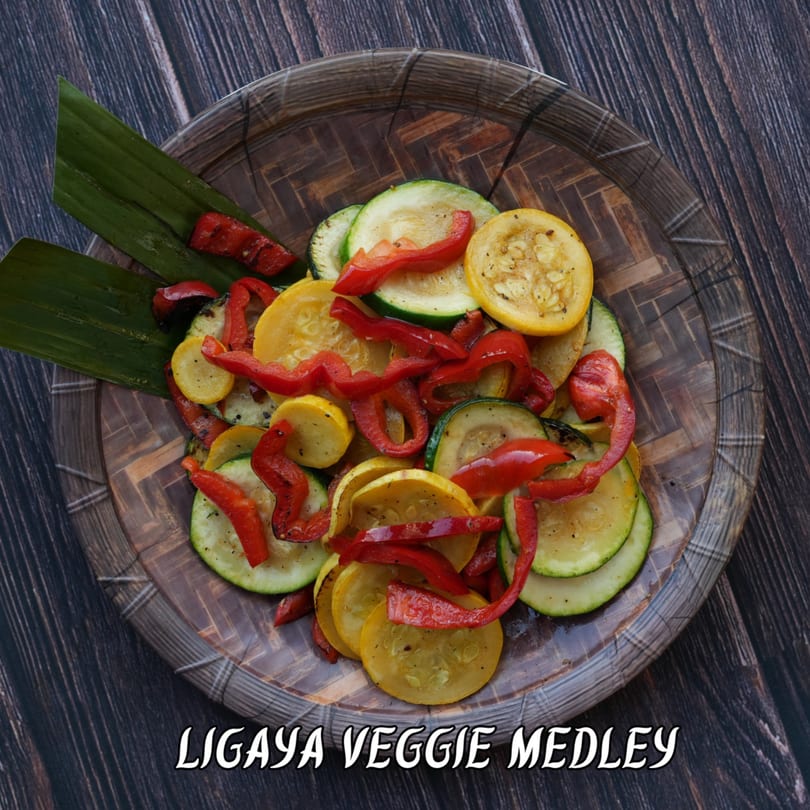 Ligaya Veggie Medley with grilled zucchini, yellow squash, and red peppers on a woven plate.