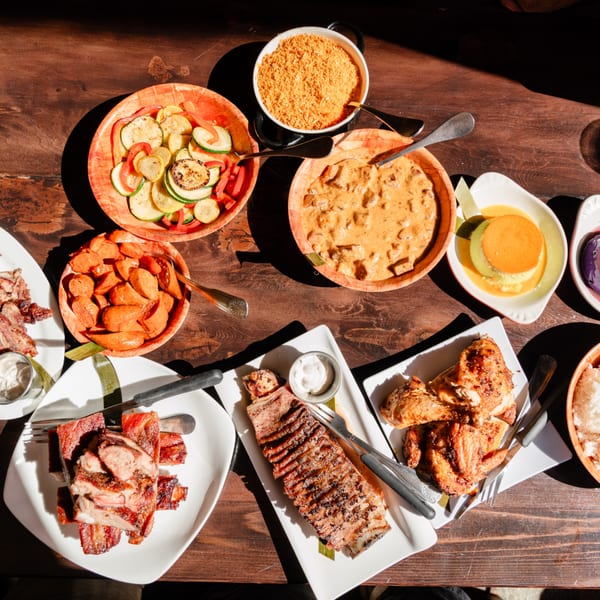 A wooden table filled with various dishes, including grilled meats, vegetables, and desserts, under bright sunlight.