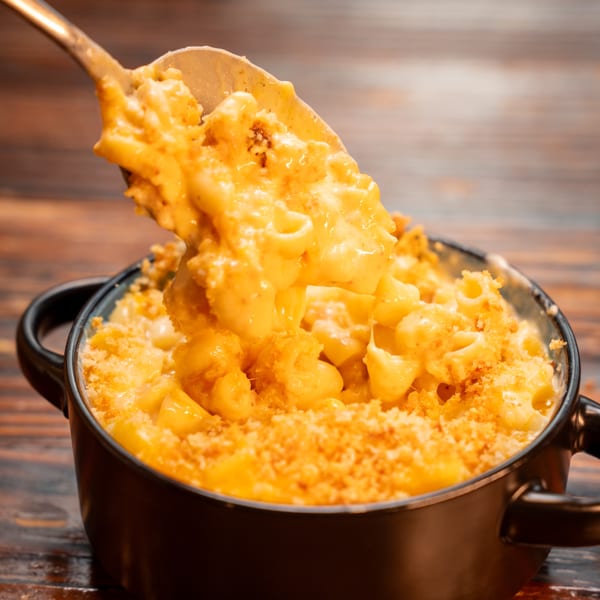 Spoon lifting creamy mac and cheese with a crispy topping from a black bowl on a wooden table.