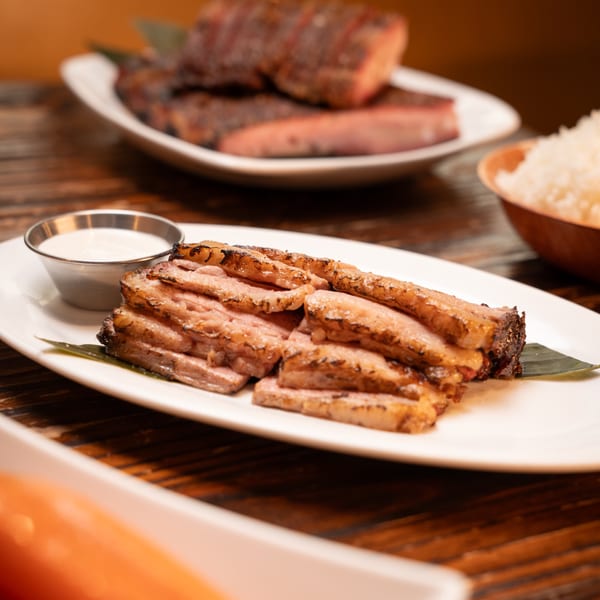 Sliced grilled pork belly with a side of white sauce, rice, and other meat dishes.