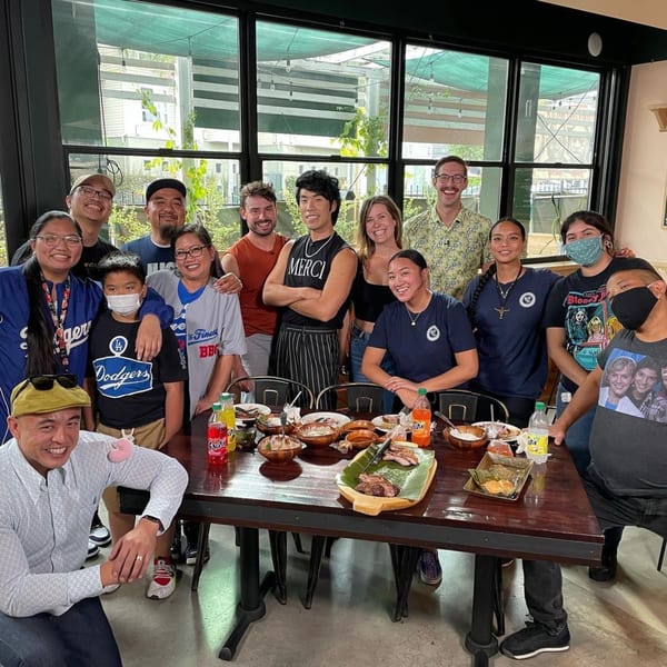 A large group of smiling people gathered around a table laden with Filipino food at The Park's Finest.